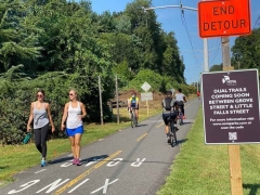 WOD-Trail-Runners Walkers and bicyclists on the W&OD Trail on a bright sunny day. Signs that say End Detour and NOVA Parks Dual Trails Coming Soon Between Grove Street and Little Falls Street. For more information visit novaparks.com or scan the QR Code.