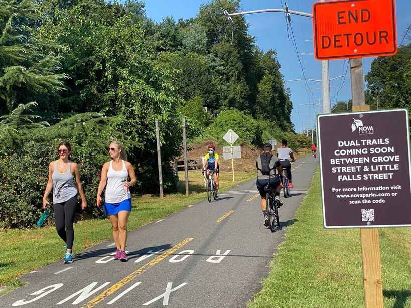 Walkers and bicyclists on the W&OD Trail on a bright sunny day. Signs that say End Detour and NOVA Parks Dual Trails Coming Soon Between Grove Street and Little Falls Street. For more information visit novaparks.com or scan the QR Code.